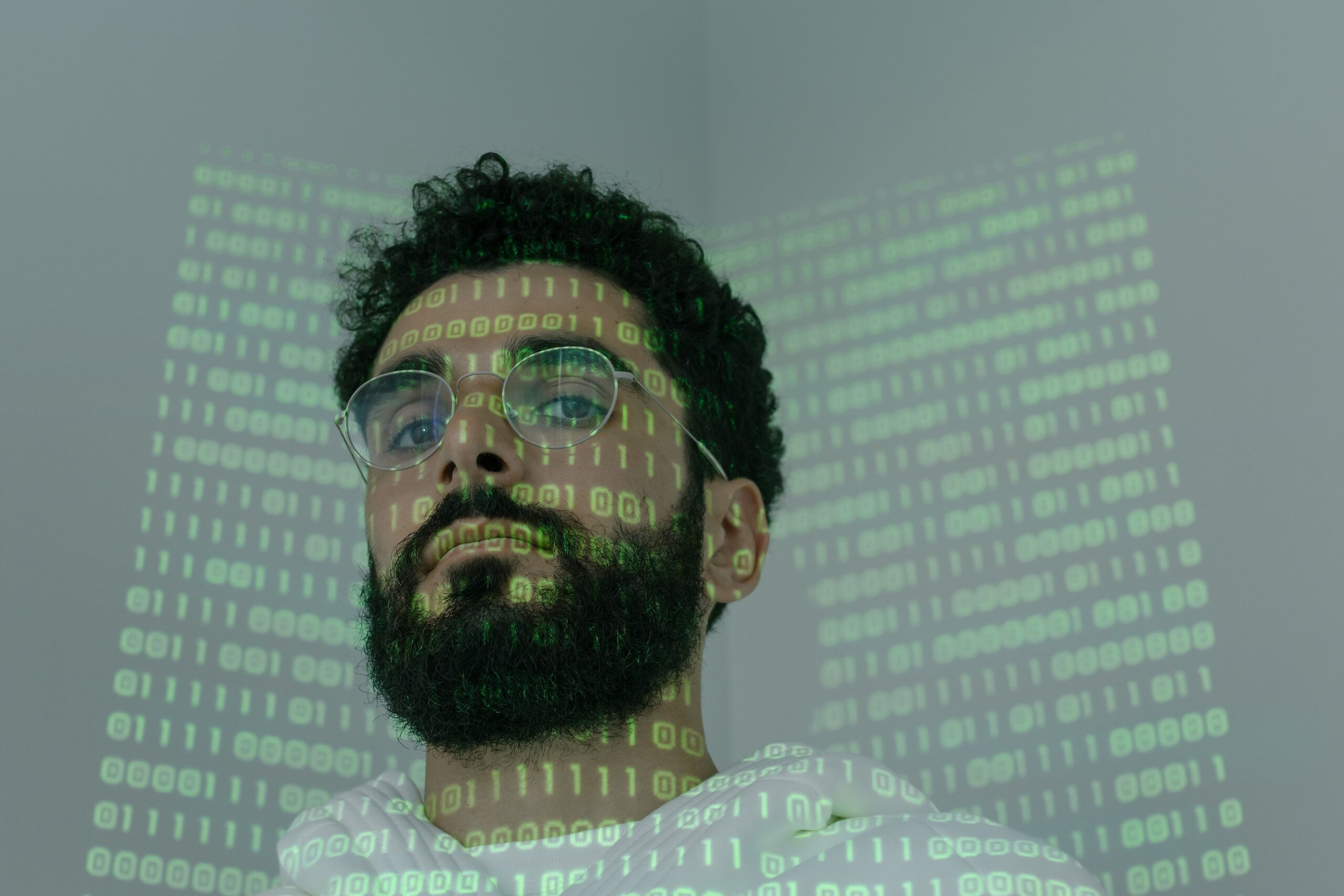 bearded, dark haired man in front of a screen with computer code, part of the code is projected on his face and head